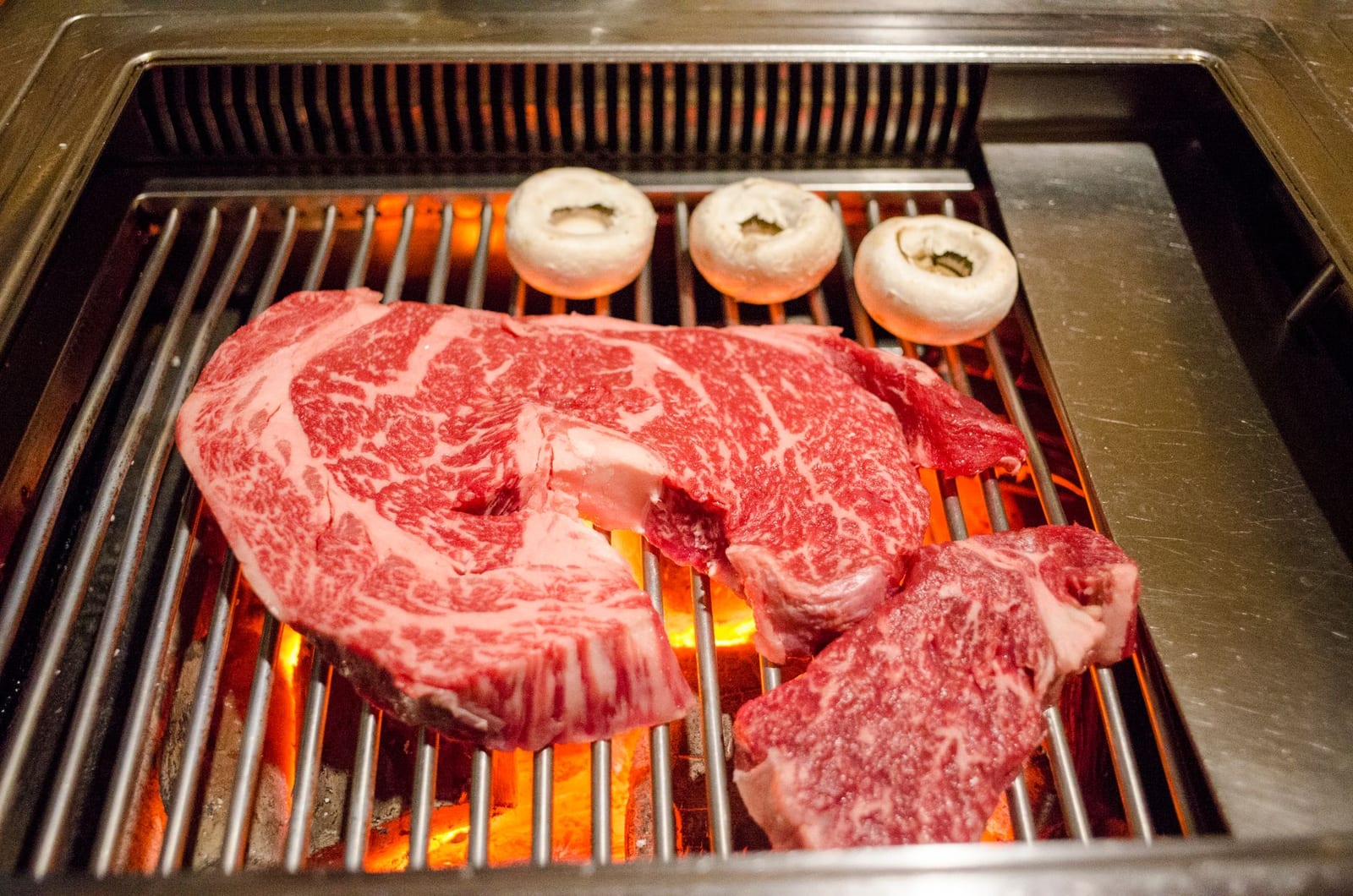 Marbled wagyu beef searing on a hot grill with mushrooms alongside